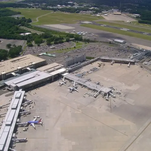High-angle aerial view of an airport terminal with several airplanes at gates, a parking garage, and a runway in the background.