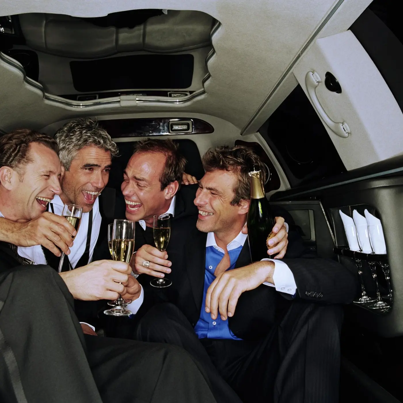 Four men in suits laughing and toasting with champagne flutes inside a luxury limousine.